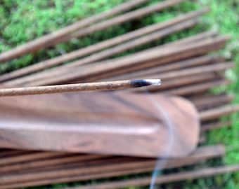 May include: A close-up of a burning incense stick with a white tip, surrounded by a bundle of other incense sticks. The incense sticks are resting on a wooden incense holder.