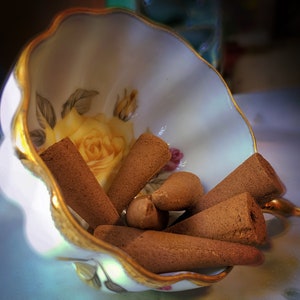Puede incluir: Una taza de t&eacute; de porcelana blanca con un borde dorado y un patr&oacute;n floral, llena de conos de incienso marrones.