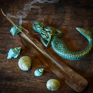 May include: A wooden incense holder with a teal mermaid figurine and seashells on a rustic wooden surface. Smoke rises from a burning incense stick.