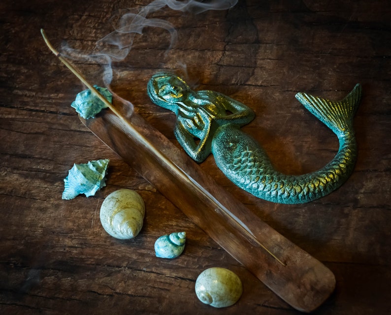 May include: A wooden incense holder with a teal mermaid figurine and seashells on a rustic wooden surface. Smoke rises from a burning incense stick.