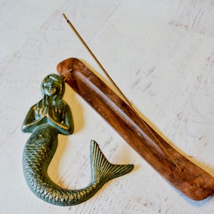 May include: A green mermaid figurine with a wooden incense holder and a stick of incense. The incense holder is brown and has a groove for the incense to sit in.