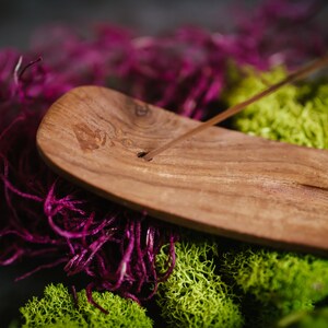 May include: A wooden incense holder with a natural wood finish. The holder is shaped like a long, narrow boat and has a groove in the center for holding incense sticks. The holder is sitting on a bed of purple and green moss.