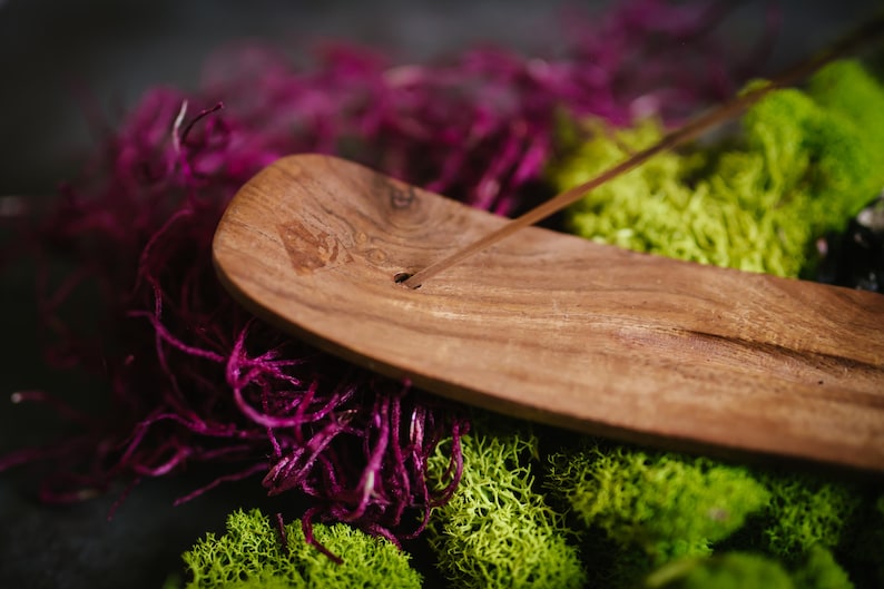 May include: A wooden incense holder with a natural wood finish. The holder is shaped like a long, narrow boat and has a groove in the center for holding incense sticks. The holder is sitting on a bed of purple and green moss.