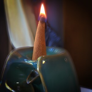 Puede incluir: Un quemador de incienso de cer&aacute;mica azul verdoso con un cono de incienso marr&oacute;n ardiendo en su interior. El cono est&aacute; encendido y tiene una peque&ntilde;a llama en la parte superior.