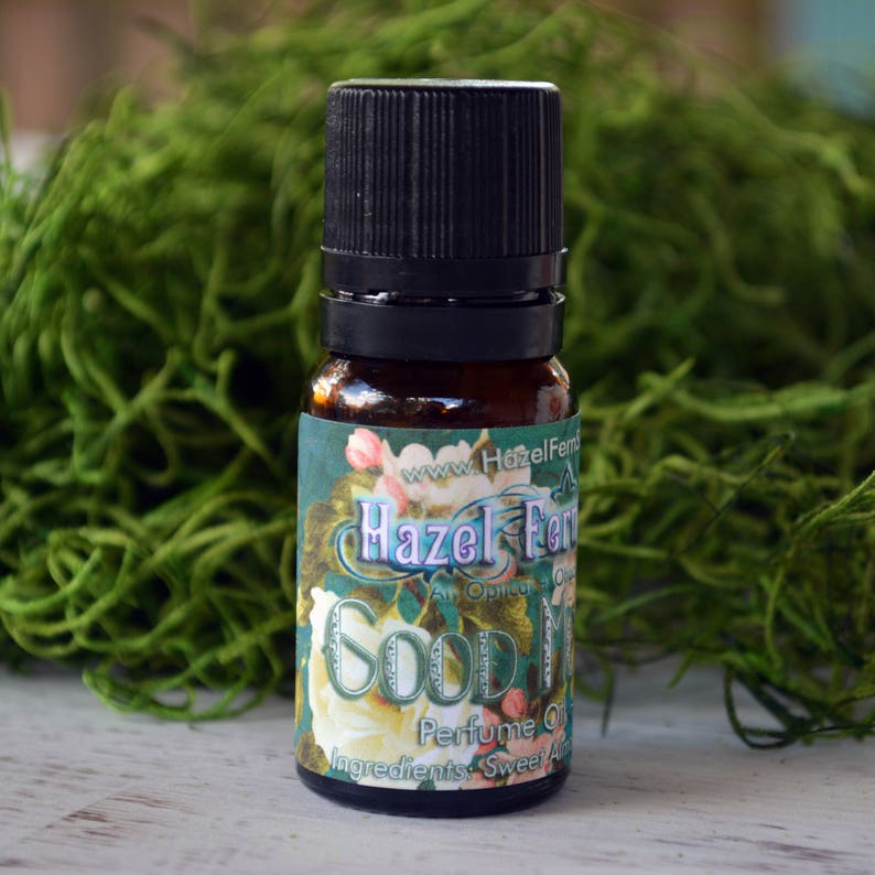 May include: A small brown glass bottle with a black cap and a label that reads "Hazel Fern An Original Oil Perfume Oil Ingredients: Sweet Almond Oil". The bottle is sitting on a white surface with green moss in the background.