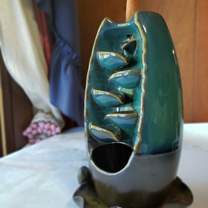 Puede incluir: Quemador de incienso de cer&aacute;mica azul verdoso y marr&oacute;n con un palo de incienso c&oacute;nico. El quemador tiene un dise&ntilde;o de cascada con varios niveles de hojas.