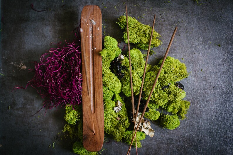 May include: A wooden incense holder with a groove down the center, surrounded by green moss and purple dried flowers. Four brown incense sticks are placed around the holder.