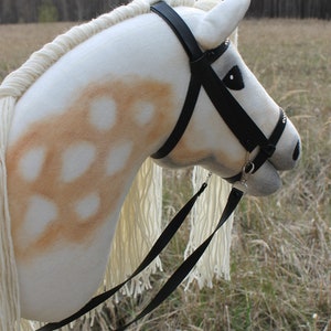 May include: A white stuffed horse head with a black leather bridle and reins. The horse has brown spots on its face and neck. The horse is standing in a field of tall grass.