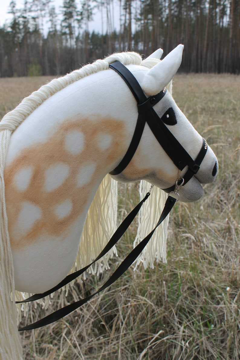 May include: A white stuffed horse head with a black leather bridle and reins. The horse has brown spots on its face and neck. The horse is standing in a field of tall grass.