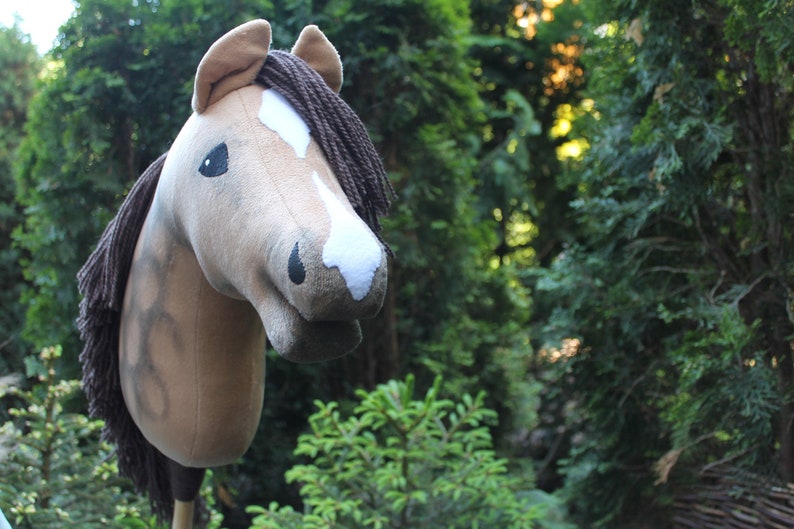 May include: A brown and white plush horse head with a brown mane, used as a hobby horse. The horse head is in focus, while the background is blurred green foliage.