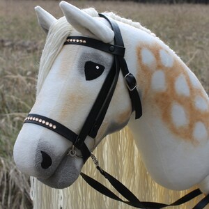 May include: A white and brown stuffed horse head with a black leather bridle and a white mane and tail. The bridle has rhinestones on the browband.