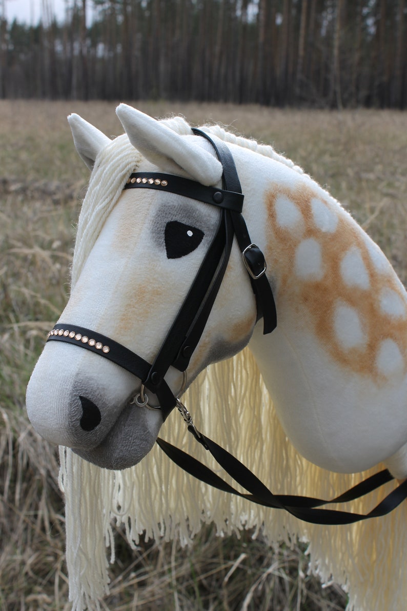 May include: A white and brown stuffed horse head with a black leather bridle and a white mane and tail. The bridle has rhinestones on the browband.