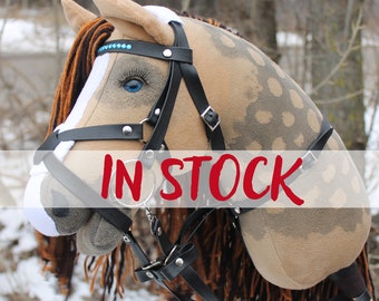 May include: A plush hobby horse head with a tan and brown spotted pattern. It features a black bridle with silver-colored hardware and blue eyes. The text "IN STOCK" is overlaid on the image. The mane and forelock are brown and black.
