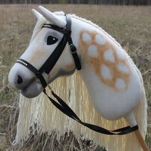 May include: A white and brown spotted hobby horse with a black leather bridle and reins. The horse has a long white mane and tail.