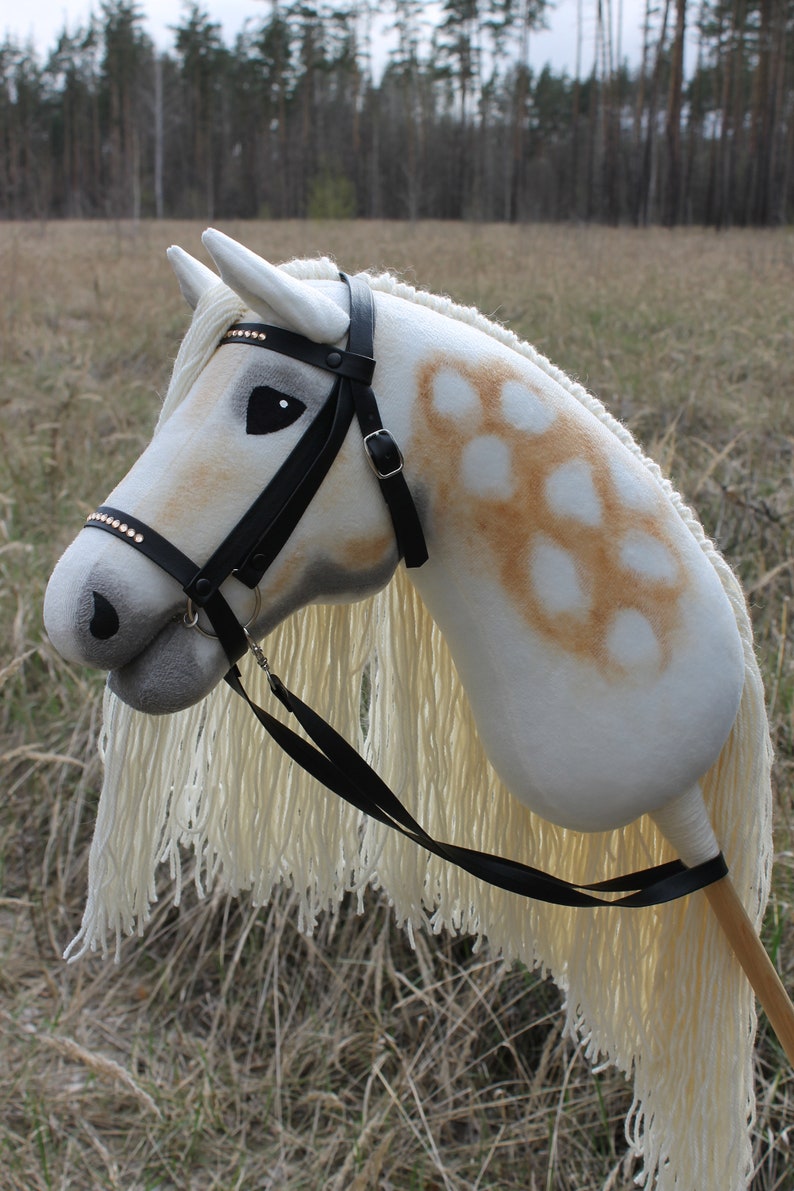 May include: A white and brown spotted hobby horse with a black leather bridle and reins. The horse has a long white mane and tail.