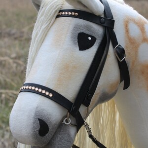 May include: A white and brown plush horse head with a black leather bridle and reins. The bridle has gold-colored rhinestones.