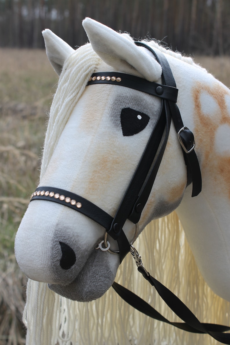 May include: A white and brown plush horse head with a black leather bridle and reins. The bridle has gold-colored rhinestones.