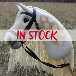 May include: A white and brown spotted hobby horse with a black bridle and reins. The horse has a white mane and tail. The hobby horse is in stock.