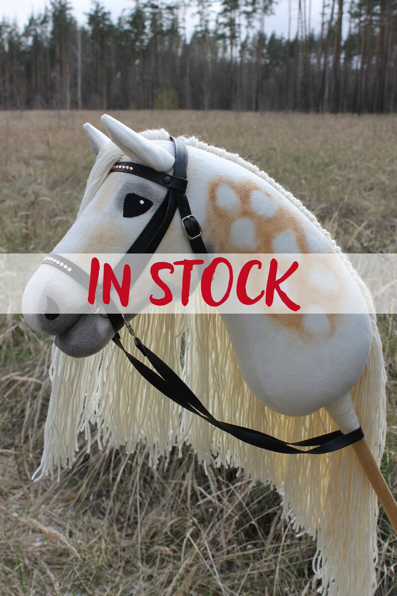 May include: A white and brown spotted hobby horse with a black bridle and reins. The horse has a white mane and tail. The hobby horse is in stock.