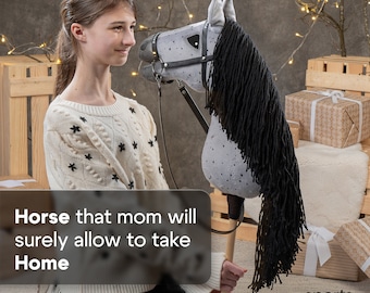 May include: A young person holds a gray and black hobby horse with a black mane and a black leather bridle. The hobby horse has a wooden stick handle. The person is wearing a white sweater with black stars. The background is a rustic wooden wall with a Christmas tree and presents.