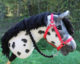 May include: A hobby horse toy featuring a white fabric head with black spots, a thick black yarn mane, and a bright red nylon bridle with silver-toned metal hardware. The horse head is attached to a smooth, light-colored wooden stick handle.