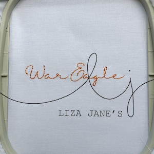 May include: A white fabric piece secured in a light gray embroidery hoop. The fabric features the orange stitched text War Eagle in a cursive font, with a black line flourish and the black printed text LIZA JANE'S positioned below it.