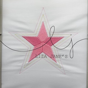 Peut inclure: Un carré de tissu blanc avec une étoile rose brodée au centre au remplissage texturé. L'étoile est entourée de deux contours cousus concentriques en fil jaune et rose. Le texte LIZA JANE'S est imprimé en noir sous l'étoile.