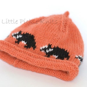 May include: A knitted orange beanie with a black and white badger design. The beanie has a folded brim and is made of soft yarn.