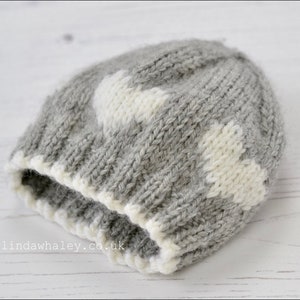 May include: Gray knitted hat with white heart designs. The hat has a white trim around the bottom edge.