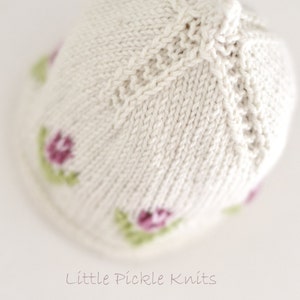 Pode incluir: Um chapéu de tricô branco para bebê com um delicado design floral. O chapéu apresenta pequenas flores cor-de-rosa com folhas verdes, criando um visual charmoso e doce. O chapéu é feito com um fio macio e confortável, perfeito para um recém-nascido.