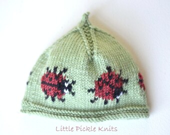 Puede incluir: Un gorro de punto verde con la parte superior puntiaguda y un borde blanco. El gorro tiene un patrón de mariquitas rojas con manchas negras.