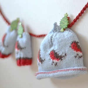 May include: A blue knitted hat and matching mittens with a red and brown robin design. The hat and mittens are hanging on a red string with a green Christmas tree clip.