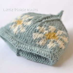 May include: A light blue knitted hat with a white and yellow daisy pattern. The hat is made by Little Pickle Knits.