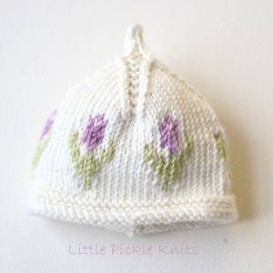 May include: White knitted baby hat with a flower design in purple and green.