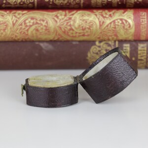 May include: A small, brown leather ring box with a hinged lid. The box is open and the inside is lined with a white fabric. The box is sitting on a white surface.