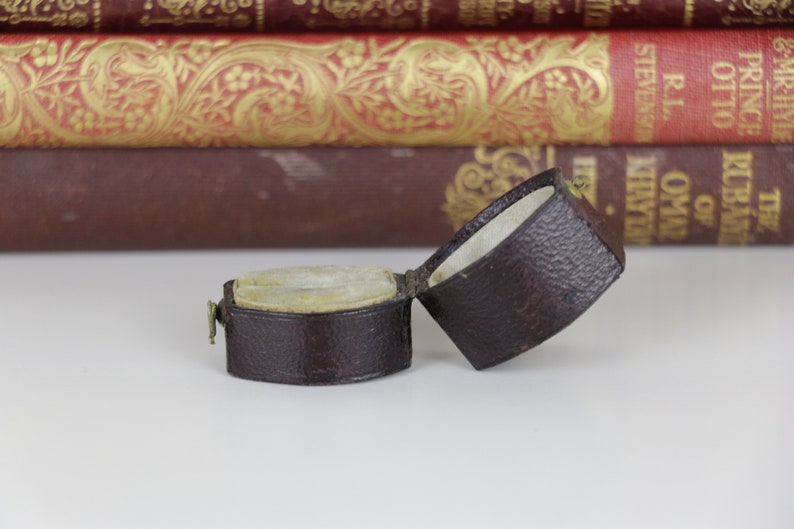 May include: A small, brown leather ring box with a hinged lid. The box is open and the inside is lined with a white fabric. The box is sitting on a white surface.