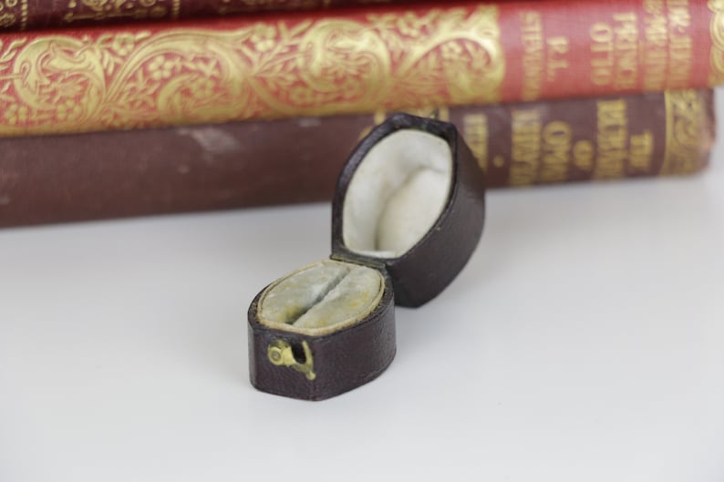 May include: A small, brown, leather ring box with a white velvet interior. The box is open and the lid is laying on its side.