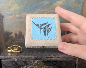 May include: A small, vintage ring box featuring a blue and white bird illustration. The box is held near a pair of gold wedding bands. The box has a light blue background with two swallows. The box is beige with gold trim.