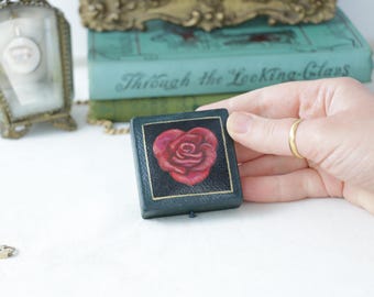 May include: A small, dark blue jewellery box featuring a red rose design on the lid. The box is rectangular with a gold border and a small clasp. The box is being held by a person.