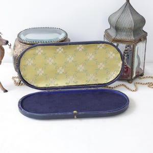 May include: An open, vintage, oval-shaped jewelry box with a yellow floral patterned interior and a dark blue velvet base. The box is set against a white background with a decorative deer figurine and a lantern.