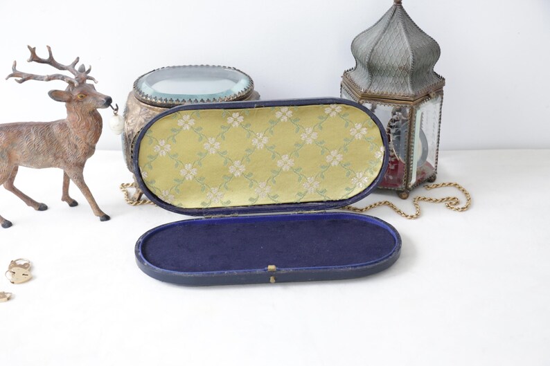 May include: An open, vintage, oval-shaped jewelry box with a yellow floral patterned interior and a dark blue velvet base. The box is set against a white background with a decorative deer figurine and a lantern.