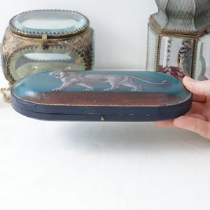 May include: An antique, oval-shaped box with a painted image of a panther. The box has a blue top with a brown base and a black frame. The box is being held by a person.