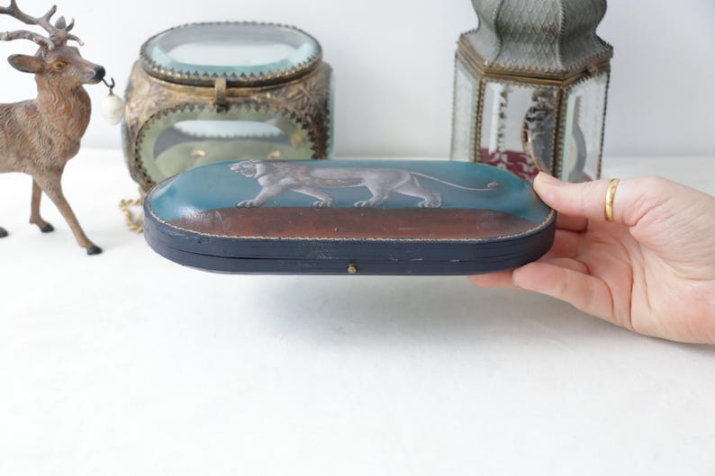 May include: An antique, oval-shaped box with a painted image of a panther. The box has a blue top with a brown base and a black frame. The box is being held by a person.
