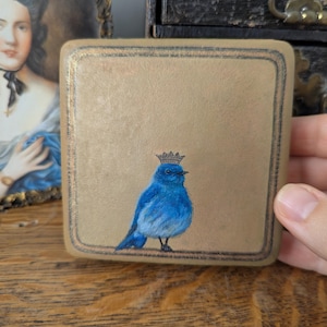 Könnte beinhalten: Eine quadratische, beigefarbene Zierbox mit einer handgemalten Illustration eines kleinen, leuchtend blauen Vogels, der eine winzige, detaillierte Krone auf dem Kopf trägt. Die Box hat eine dünne, dunkelbraune Umrandung um das Bild.