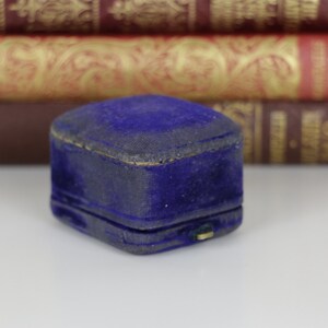 May include: A small, vintage, blue velvet jewelry box. The box is rectangular with a hinged lid and a gold-colored clasp. The velvet has a slightly worn appearance. The background features a stack of antique books.