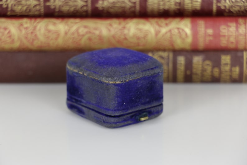 May include: A small, vintage, blue velvet jewelry box. The box is rectangular with a hinged lid and a gold-colored clasp. The velvet has a slightly worn appearance. The background features a stack of antique books.