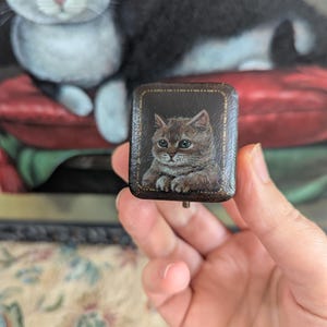May include: A small, square, dark brown box featuring a detailed, hand-painted portrait of a brown tabby cat on its lid. The lid is bordered by a thin, gold-coloured line, and the box has a smooth, polished finish with a small metal latch visible.