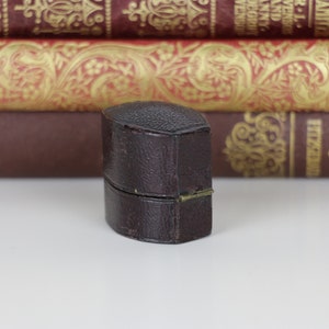 May include: A small, dark brown, leather-bound box with a hinged lid. The box is closed and sitting on a white surface.