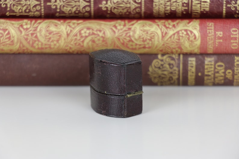 May include: A small, dark brown, leather-bound box with a hinged lid. The box is closed and sitting on a white surface.