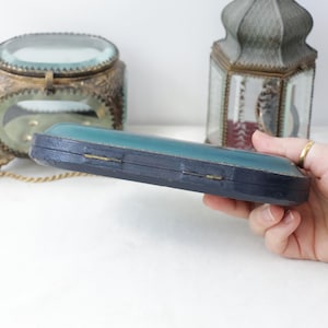 May include: A vintage, rectangular jewelry box with a teal top and dark blue sides. The box is held in a hand, with other antique decorative items in the background, including a deer figurine and a glass-covered container.